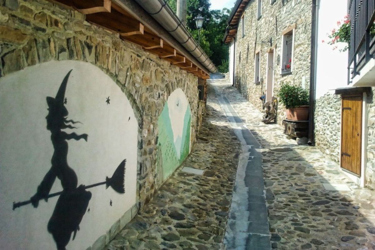 Vicolo borgo medievale in pietra Italia, murales strega scopa profilo nero, atmosfera magica halloween, destinazione misteriosa borgo streghe