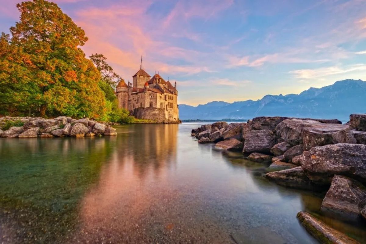 Castello di Chillon sulle rive di un lago al tramonto, circondato da rocce e montagne.